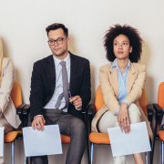 Imagem de várias pessoas sentadas em uma fila com currículos na mão, representando a busca por um trabalho em um cenário de desemprego estrutural.