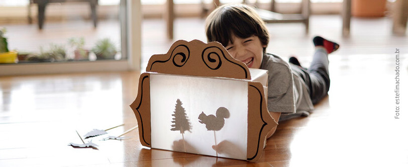 criança deitada no chão brincando com uma caixa de sapatos transformada em teatrinho de sombras