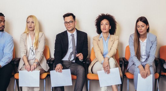 Imagem de várias pessoas sentadas em uma fila com currículos na mão, representando a busca por um trabalho em um cenário de desemprego estrutural.