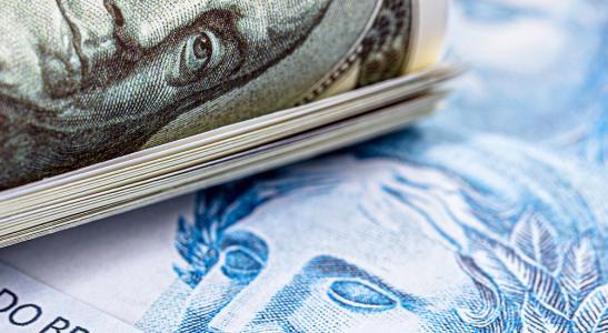 vários dólares enrolados por uma liga acima de uma base composta por notas de 100 reais, simbolizando como investir em dólar no Brasil.